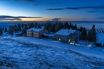 Domžalski dom mountain lodge on Velika planina, illuminated during the blue hour at dawn, set against the snow-covered plateau and pine trees, with fog filling the valley below
