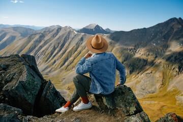 Young hispanic male hiker in denim and hat relaxing on mountain cliff overlooking scenic landscape, back view