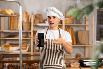 Portrait of friendly male baker with mobile phone in hands in bakery interior