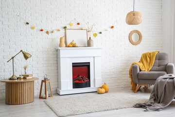 Fototapeta premium Interior of stylish room with modern fireplace, armchair, table and autumn decorations near white brick wall