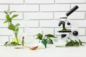 Laboratory glassware and microscope with ingredients for preparing natural cosmetics on white table near brick wall