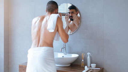 A man stands in a contemporary bathroom, towel around his waist, using a mirror to groom himself. Natural light streams in, creating a fresh atmosphere for his morning routine.