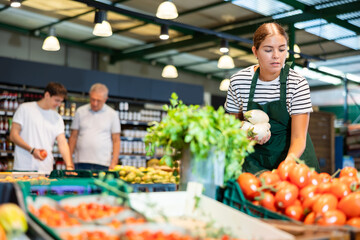 Focused skilled young saleswoman in green apron working in fruit and vegetable section of supermarket, laying out white eggplants on display stand