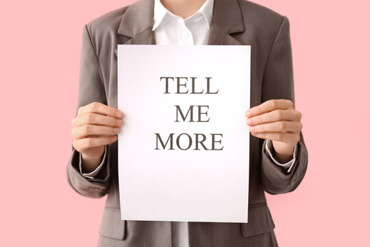 Businesswoman holding paper with text TELL ME MORE on pink background, closeup. Gossip concept