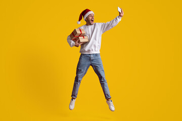 A man wearing a Santa hat jumps happily while holding wrapped gifts and taking a selfie. The bright yellow background adds to the joyful festive atmosphere.