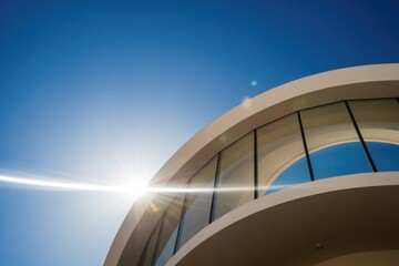 Modern curved architecture with glass windows under a bright blue sky. Sun's lens flare streaks across the contemporary building facade.