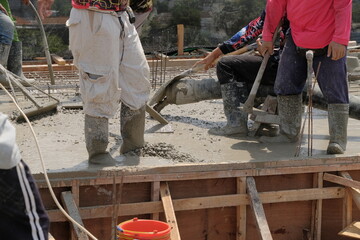 construction workers in boots using a large pipe and pump to pour wet, fluid concrete onto a floor slab at a building site