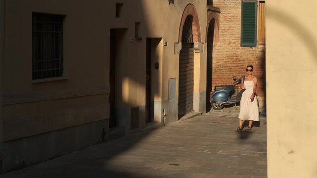 Tourist walking in a typical italian street. Young woman wearing sunglasses and white dress strolling through narrow cobblestone street in italian town, scooter parked alongside stone buildings