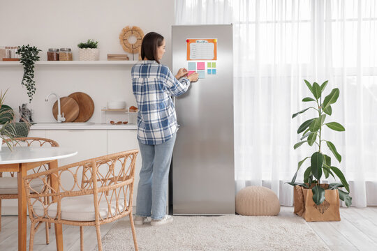 Young woman attaching sticky notes on fridge in modern kitchen - Powered by Adobe