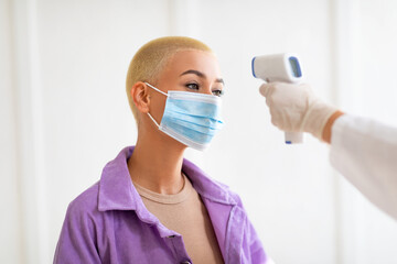 Doctor measuring woman's temperature with electronic thermometer before covid vaccination at clinic. Young lady in face mask participating in coronavirus immunization campaign