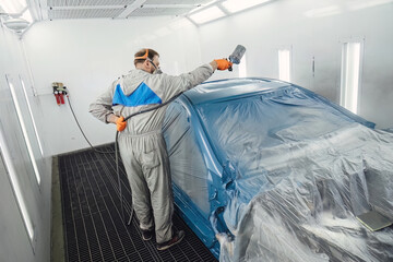 Professional painter in a protective suit spraying paint onto the car body inside a clean, modern spray booth. High quality automotive repair and bodywork service maintenance for vehicles.