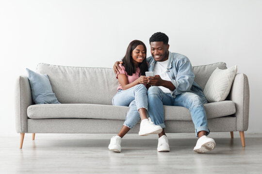 African American couple sharing social media on cellphone, happy partners sitting on the sofa, male showing gadget. Smiling young black guy embracing lady looking at smartphone screen, free copy space