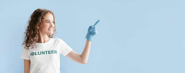 A young woman with curly hair is wearing a white volunteer shirt and blue gloves. She smiles and points with her gloved hand, conveying a positive message in a bright blue setting.
