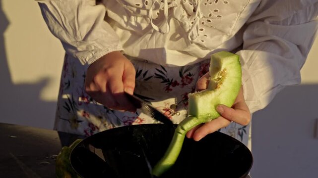 Detailed close-up of a person's hands skillfully peeling a green melon with a knife. The process reveals fresh fruit, destined for a bowl on the kitchen table.