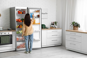 Young woman taking bottle of water from open fridge full of food in kitchen, back view