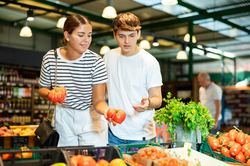 Cheerful young couple, girl and guy, shopping in farm store, choosing ripe red tomatoes..