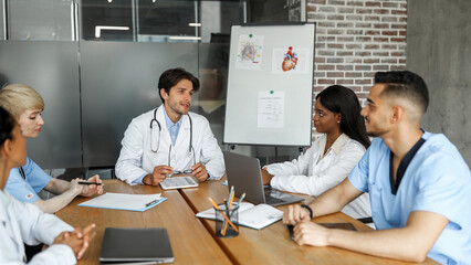 Handsome young man chief physician having breefing with his male and female international colleagues, dicsussing difficult situations while COVID-19 pandemic, panorama with copy space