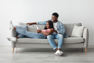 African American couple sharing social media on cellphone, happy partners sitting on the sofa, female lying on guy's lap and showing gadget. Smiling young man and woman looking at smartphone screen