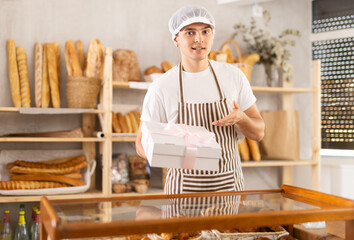 Smiling young salesman in striped apron presenting made-to-order cake, elegantly packaged in white gift box with pink ribbon, in small craft bakery