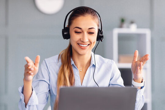 A young woman with long hair wearing a light blue shirt and headphones smiles brightly while participating in an online meeting. Her body language shows engagement as she gestures with her hands.