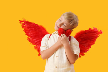 Obraz premium Cute little happy boy dressed as cupid with knitted heart toy on yellow background. Valentine's Day celebration