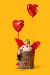 Obraz premium Cute little happy boy dressed as cupid in wicker basket with heart-shaped balloons and binoculars on yellow background. Valentine's Day celebration