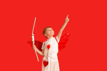 Obraz premium Cute little happy boy dressed as cupid with bow and arrow pointing at something on red background. Valentine's Day celebration