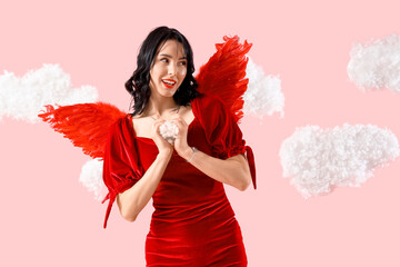 Obraz premium Beautiful young happy woman dressed as cupid in clouds showing heart gesture on pink background. Valentine's Day celebration