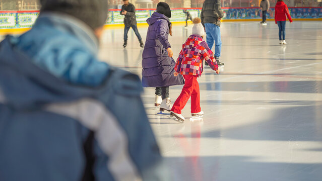 People ice skating on open-air rink during sunny winter day, wearing colorful jackets and warm clothes. Concept of active winter leisure, joyful family ice skating experience