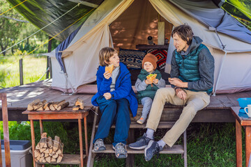 Father with his toddler and teenage son tourists in warm vests and hats eating buns while sitting on glamping steps, enjoying family travel, outdoor adventure, and bonding