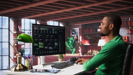 African american man looks at notes on terminal window for visual programming, typing code on software at his home office. Coding application UI used by engineer writing and testing code. Camera B.