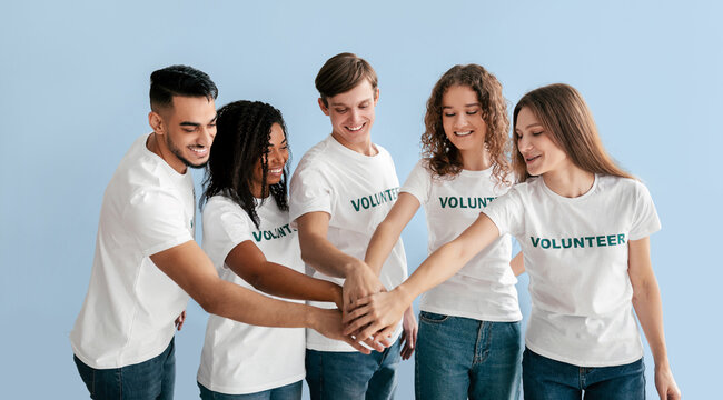 A diverse group of young adults stands in a light blue setting, wearing white t-shirts with the word 'VOLUNTEER' printed on them.