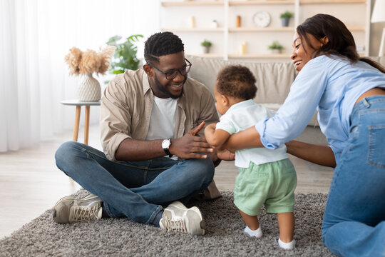 A joyful black toddler takes his first steps in the living room while his mother supports him. His father sits on the floor, smiling, as they share a special moment in their family space.