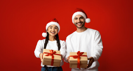 A joyful couple stands against a bright red background wearing matching Santa hats. They hold beautifully wrapped presents with ribbons. The scene captures the excitement of the holiday season.