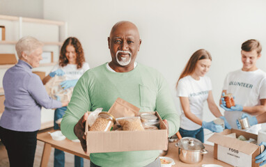 A group of volunteers works together, packing food items for distribution. One man holds a box filled with various food products, while others assist in the background.