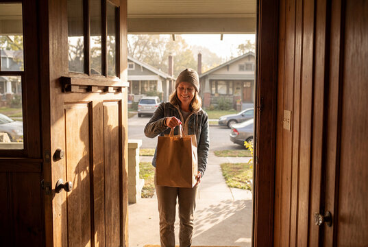 Female delivery driver handing grocery bag to customer at front door for home delivery service.