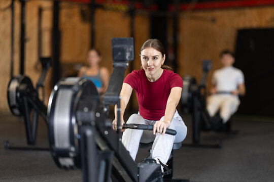 Girl hardly performs exercises on rowing machine during fitness class. Gym and fitness equipment, visitors are engaged in physical exercises during training