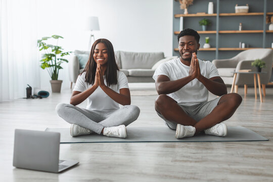 Smiling black couple meditating looking at pc screen sitting in lotus pose asana on floor holding hands palms together in prayer position, watching online video tutorial, guy and lady training at home - Powered by Adobe