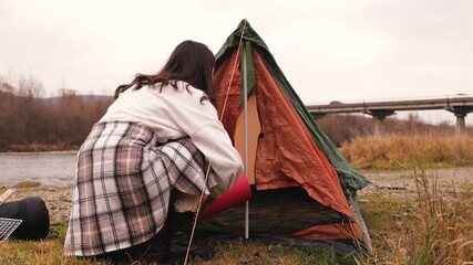 Woman setting up her camping tent by placing a sleeping bag and sleeping mat inside during an outdoor trip near the river, preparing the interior for a comfortable overnight stay