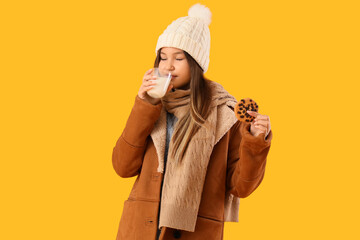 Cute teenage girl in warm clothes with delicious cookie and glass of milk on yellow background