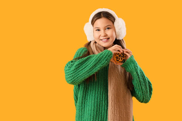 Cute teenage girl in warm clothes with delicious cookie making heart gesture on yellow background