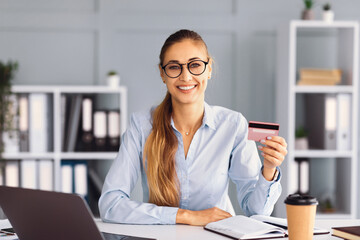 A professional woman is smiling while holding a credit card in her hand. She sits at a modern work desk surrounded by a laptop, notebook, and a cup of coffee, indicating a productive atmosphere.