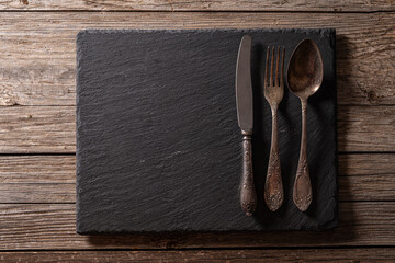 Retro metal cutlery on granite surface over aged wooden background. Vintage cutlery on granite serving board over rustic wood. Old-Style fork knife and spoon on granite plate.