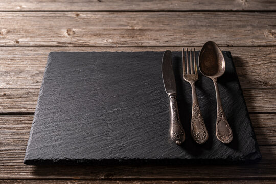 Retro metal cutlery on granite surface over aged wooden background. Vintage cutlery on granite serving board over rustic wood. Old-Style fork knife and spoon on granite plate. - Powered by Adobe