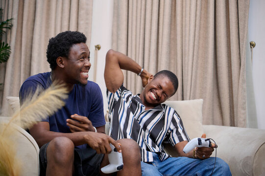 Two young Black men sit on a couch indoors, laughing together while holding video game controllers during a playful gaming moment.