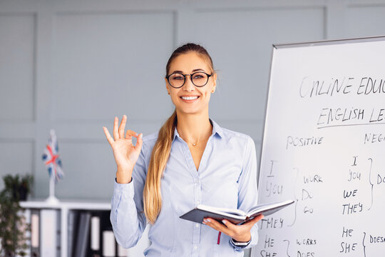 A confident teacher stands in a modern classroom, showing a thumbs up gesture while holding a notebook.