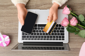 Female hands with mobile phone, credit card, pink roses and modern laptop on wooden background. Valentine's Day celebration