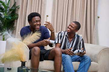 Two young African men sit on a couch laughing together while holding game controllers during a relaxed gaming session.
