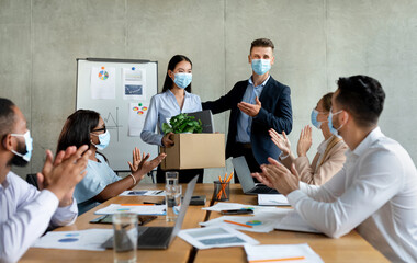 In a modern conference room, diverse coworkers in medical face masks applaud as a new team member is welcomed with a box and a plant. Excitement fills the air during this corporate gathering.