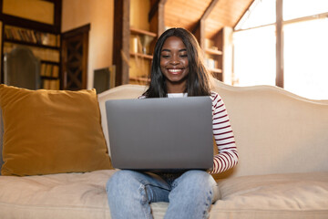 Naklejka premium A young woman with long, dark hair is smiling as she types on a laptop while sitting comfortably on a sofa. The room is bright with ample natural light and cozy decor.